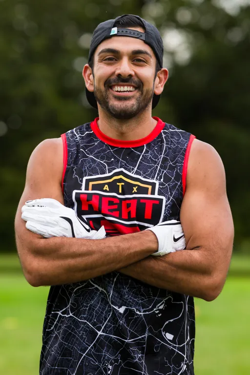 An ATX Heat player stands on a grassy practice field for Gay Bowl 2025, smiling with arms crossed. The athlete wears a black-and-red sleeveless ATX Heat jersey, white receiver gloves, and a backward cap, with trees blurred in the background.