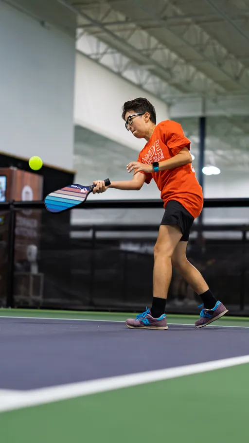 A player in a bright orange "Pride Pickleball" shirt and black shorts is actively hitting a pickleball with a paddle on an indoor court. The athlete is wearing glasses and athletic shoes, and is focused on returning the ball mid-play.