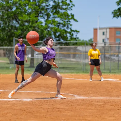 A player in a purple tank top and shorts is in mid-action, preparing to throw a kickball on a dirt field, while other competitors wearing purple and yellow shirts stand ready in the background. The setting is outdoors with a chain-link fence, trees, and a building visible beyond the field.