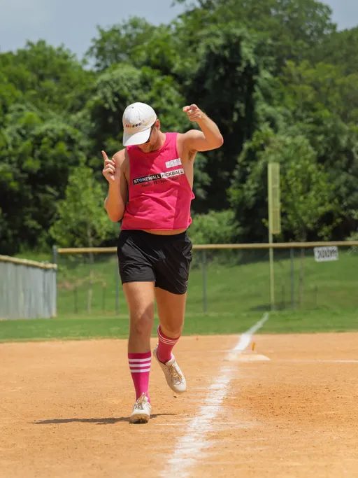 A player wearing a red "Stonewall Kickball" jersey, black shorts, pink striped socks, white sneakers, and a white cap is running along the baseline on a sandy kickball field, raising both arms in a celebratory gesture. Trees and a fence are visible in the background.