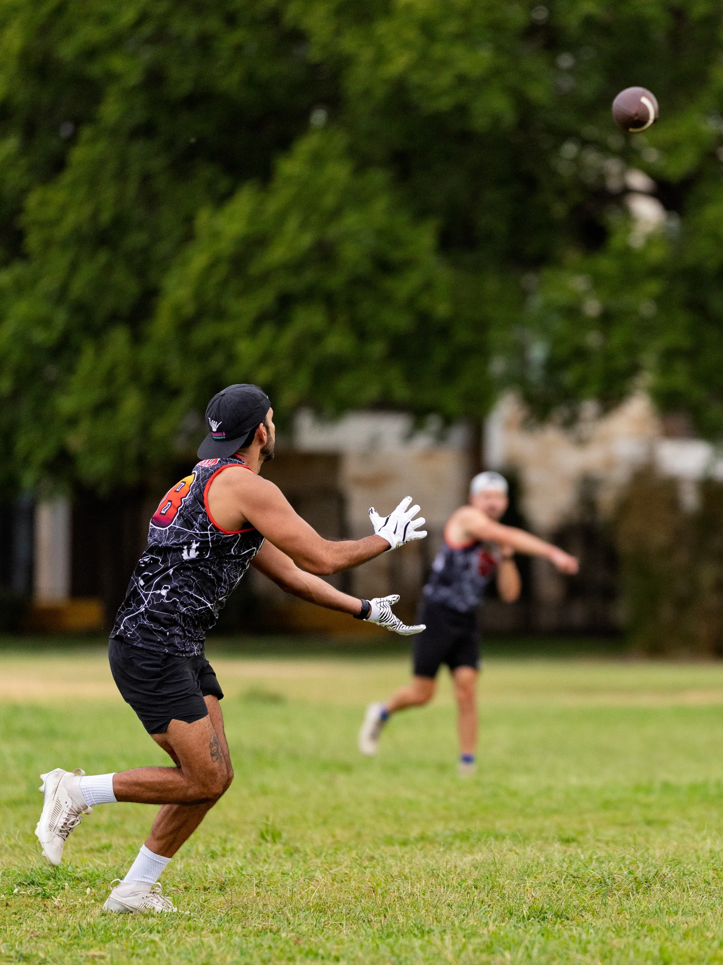 On a grassy park field, an ATX Heat (Orange) receiver in a black-and-orange sleeveless jersey, white gloves, and cleats tracks a pass flying toward them. In the background, a teammate in matching practice gear completes the throw, with trees and buildings blurred—capturing a passing drill for Gay Bowl 2025 prep.