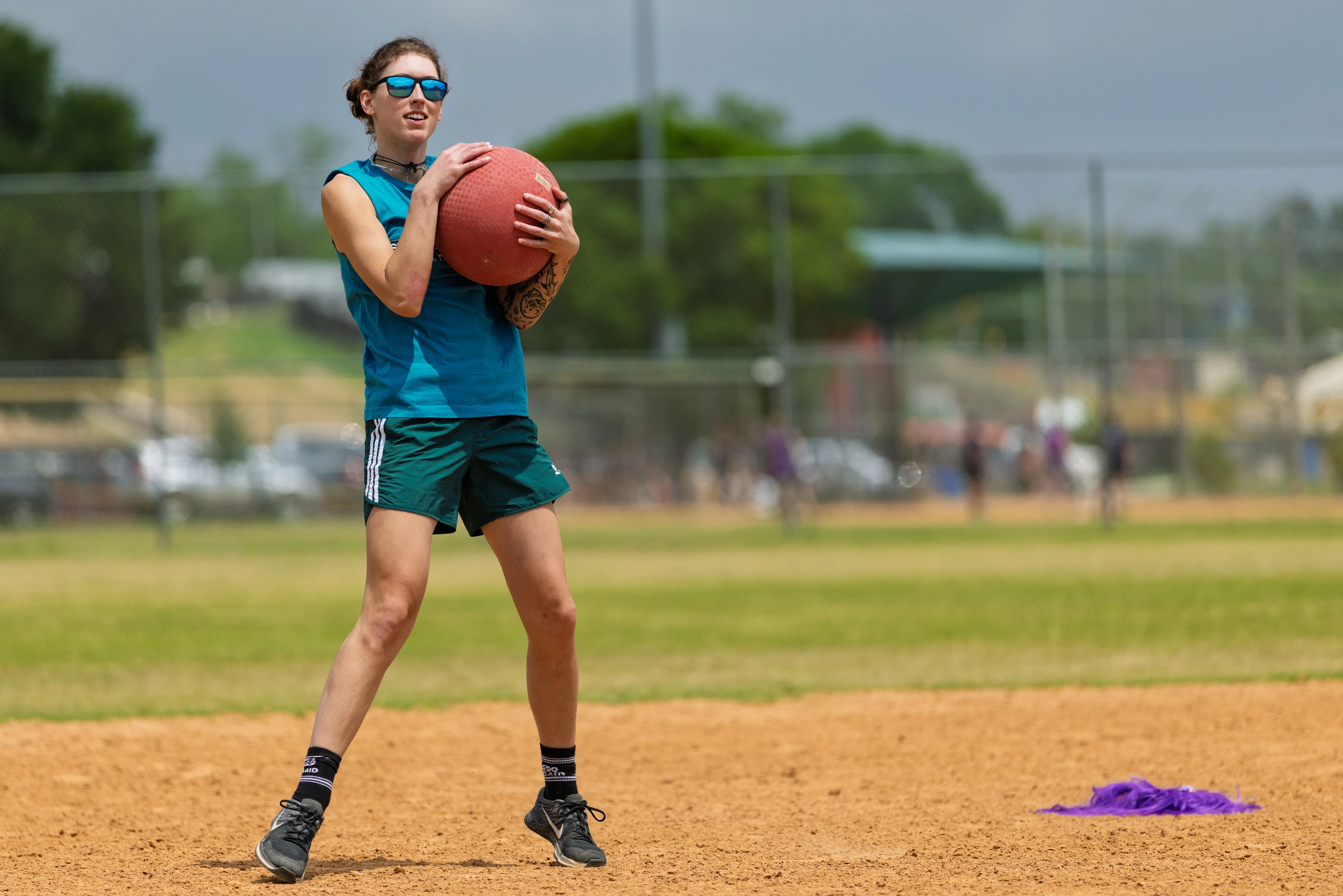A player wearing sunglasses, a sleeveless blue shirt, and athletic shorts stands on a kickball field holding a red kickball, appearing ready to throw or pitch; a purple cloth lies on the dirt nearby and a chain-link fence is visible in the background.