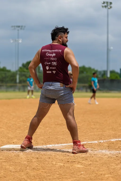 A kickball player stands at a base on a sandy field, wearing a sleeveless maroon shirt with sponsor logos, denim shorts, and red shoes, while other players and the outfield are visible in the background.