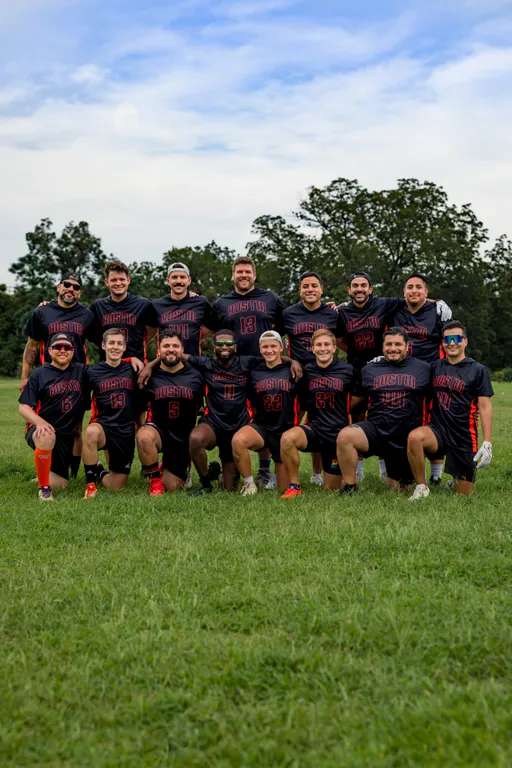 ATX Heat (Orange) flag football team poses in two rows on a grassy field, some kneeling and some standing, smiling for a group shot. The players wear black uniforms with orange accents and “Austin” lettering with numbers, cleats, and occasional accessories like caps and sunglasses, with trees and an overcast sky in the background.