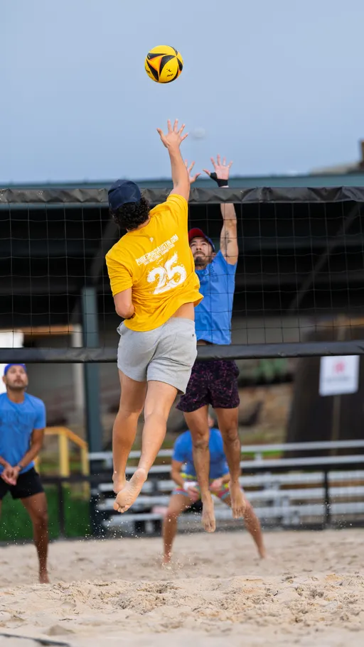 A volleyball player in a yellow shirt and gray shorts jumps to hit the ball over the net, while an opponent in a blue shirt leaps with arms extended, attempting to block the shot on a sandy outdoor volleyball court.