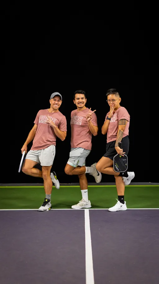 Three pickleball players strike playful poses at the center of a court, all wearing matching "Pride Pickleball" shirts, athletic shorts, and sneakers, with one holding a pickleball paddle. The background is a dark indoor setting, highlighting the players and the court lines beneath them.