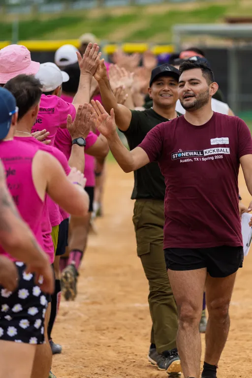 Players wearing team shirts, including one in a maroon "Stonewall Kickball Austin, TX | Spring 2025" shirt, are giving high-fives in two lines on a kickball field, celebrating after a game. The setting is an outdoor dirt field, and the players are dressed in athletic clothing, with some wearing hats and shorts.