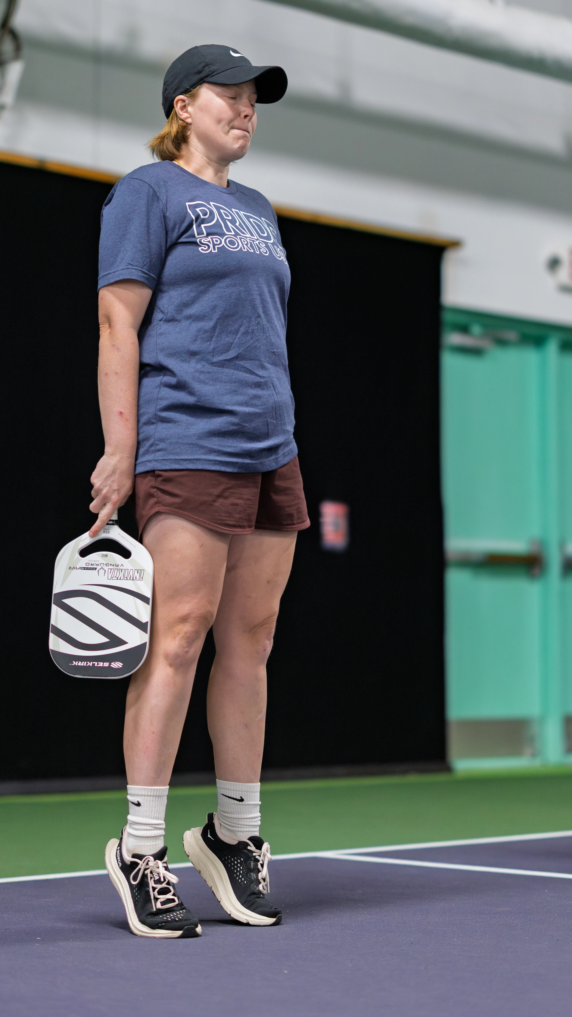 A pickleball player stands on an indoor court, holding a paddle at their side, wearing a "Pride Sports" t-shirt, athletic shorts, a cap, and sneakers. The player appears to be focused, possibly waiting for their turn during a game or practice session.