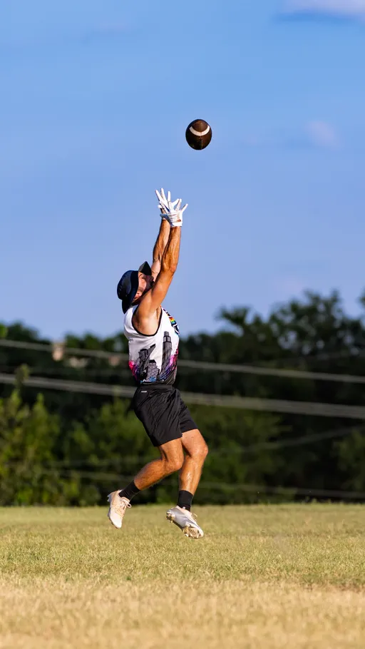 ATX Heat (Black) player leaps on a grassy field during flag football practice, arms extended in white receiver gloves to catch a football, wearing a black cap, white tank with a colorful skyline graphic, black shorts, and white cleats, with trees and power lines in the background under a clear blue sky.