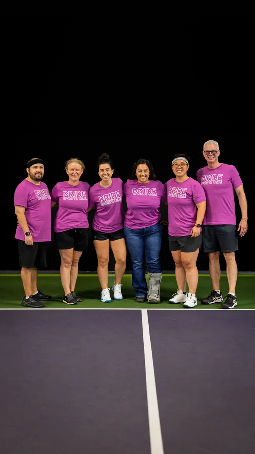 A group of six players stands together on a pickleball court wearing matching bright pink “Pride Pickleball” shirts, with some in athletic shorts and sneakers and one person wearing a medical boot.