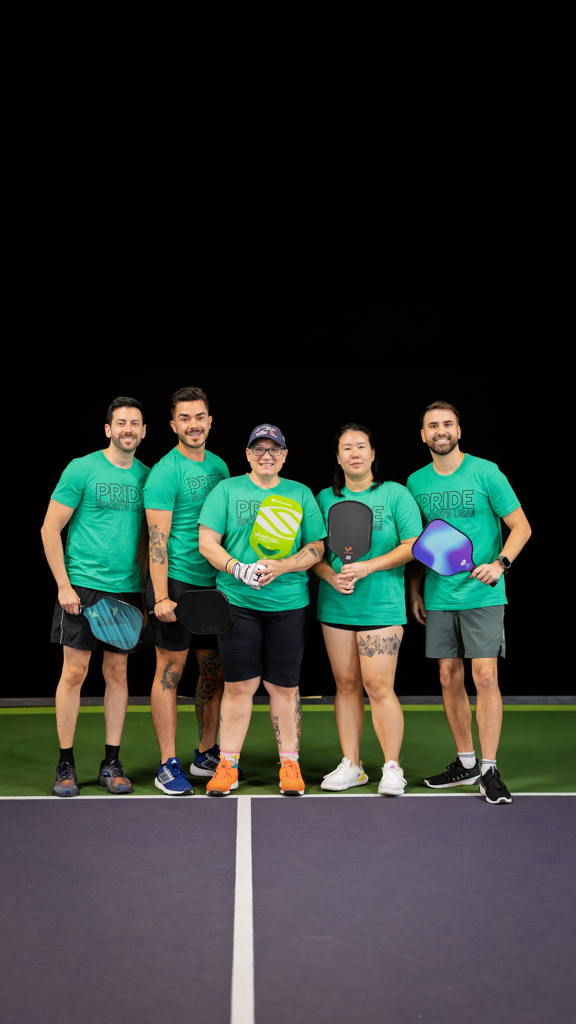 Five pickleball players stand together on an indoor court, each holding a pickleball paddle and wearing matching green "Pride" t-shirts, athletic shorts, and sneakers. The group is positioned behind the net, smiling and appearing ready for a game during the Pride Pickleball Summer 2025 season.