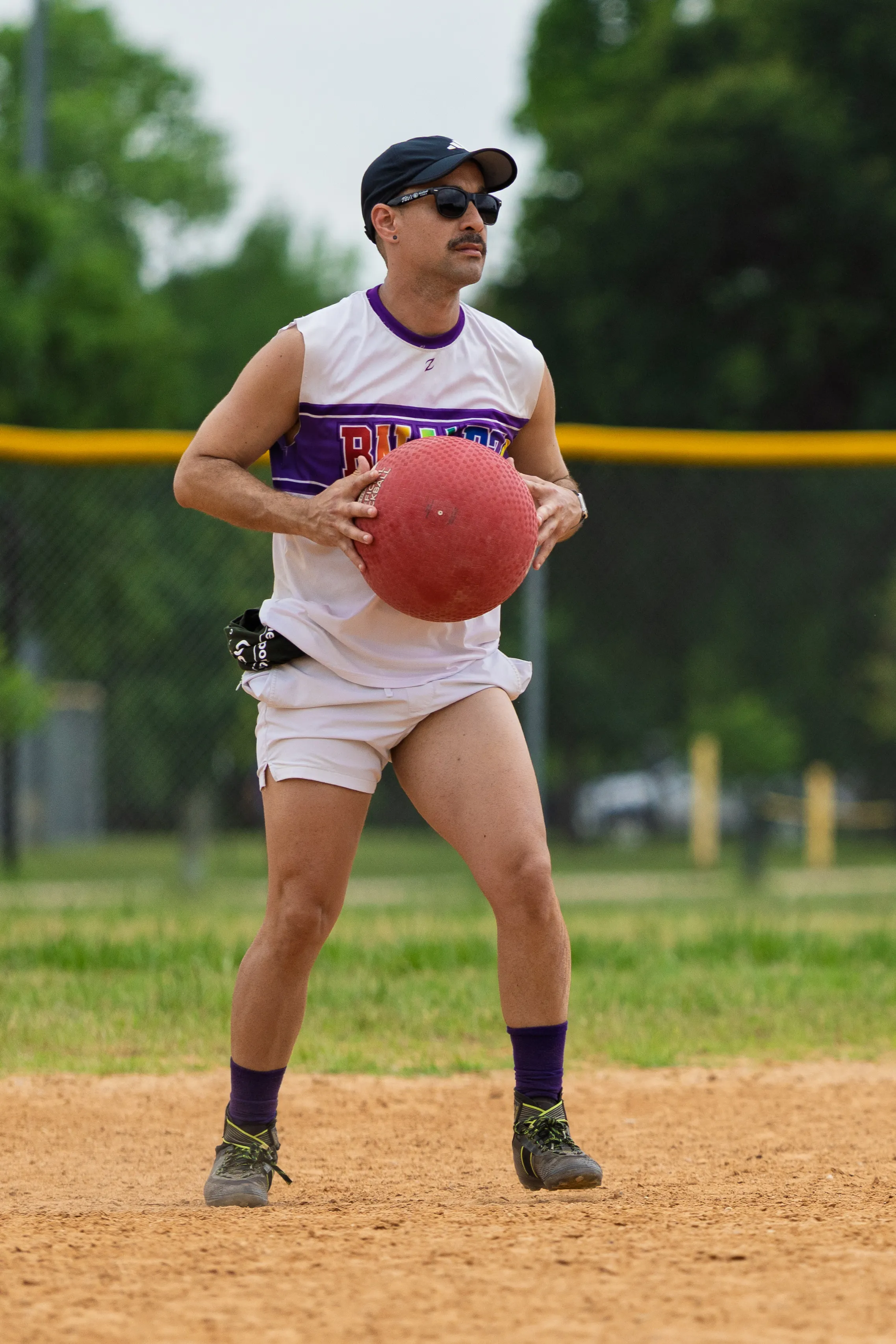On a sandy kickball infield, a player in a white-and-purple jersey and shorts grips a red kickball with both hands, pausing in a ready stance as if preparing to throw to a base. The player wears a black cap, sunglasses, purple socks, and cleats, with a yellow outfield fence and trees in the background.