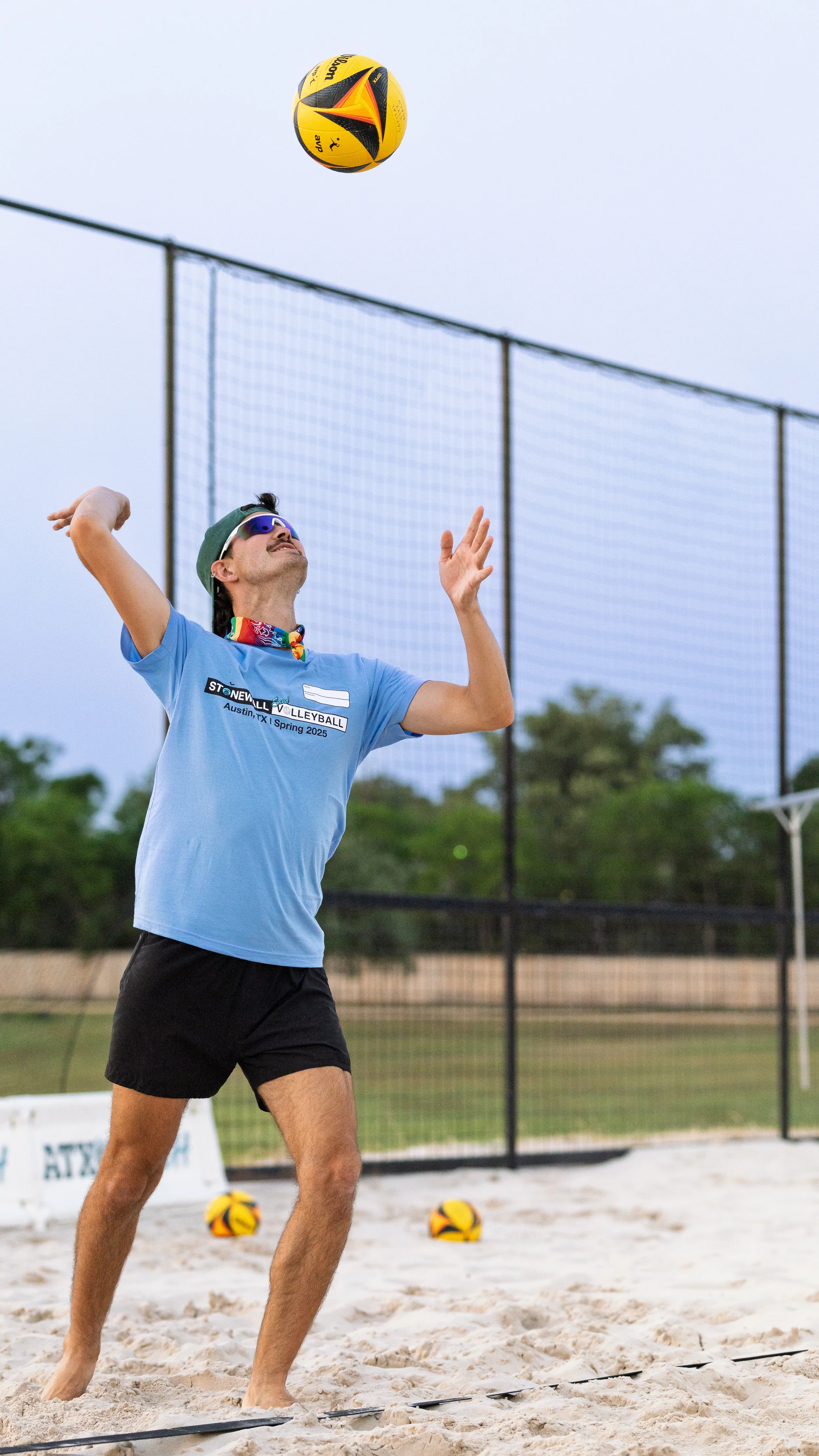A player on a sand volleyball court tosses a yellow-and-black ball overhead to serve, wearing a light-blue Stonewall Volleyball Spring 2025 shirt, black shorts, sunglasses, a backward cap, and a rainbow bandana. Extra balls rest in the sand, with tall perimeter netting and trees in the background.