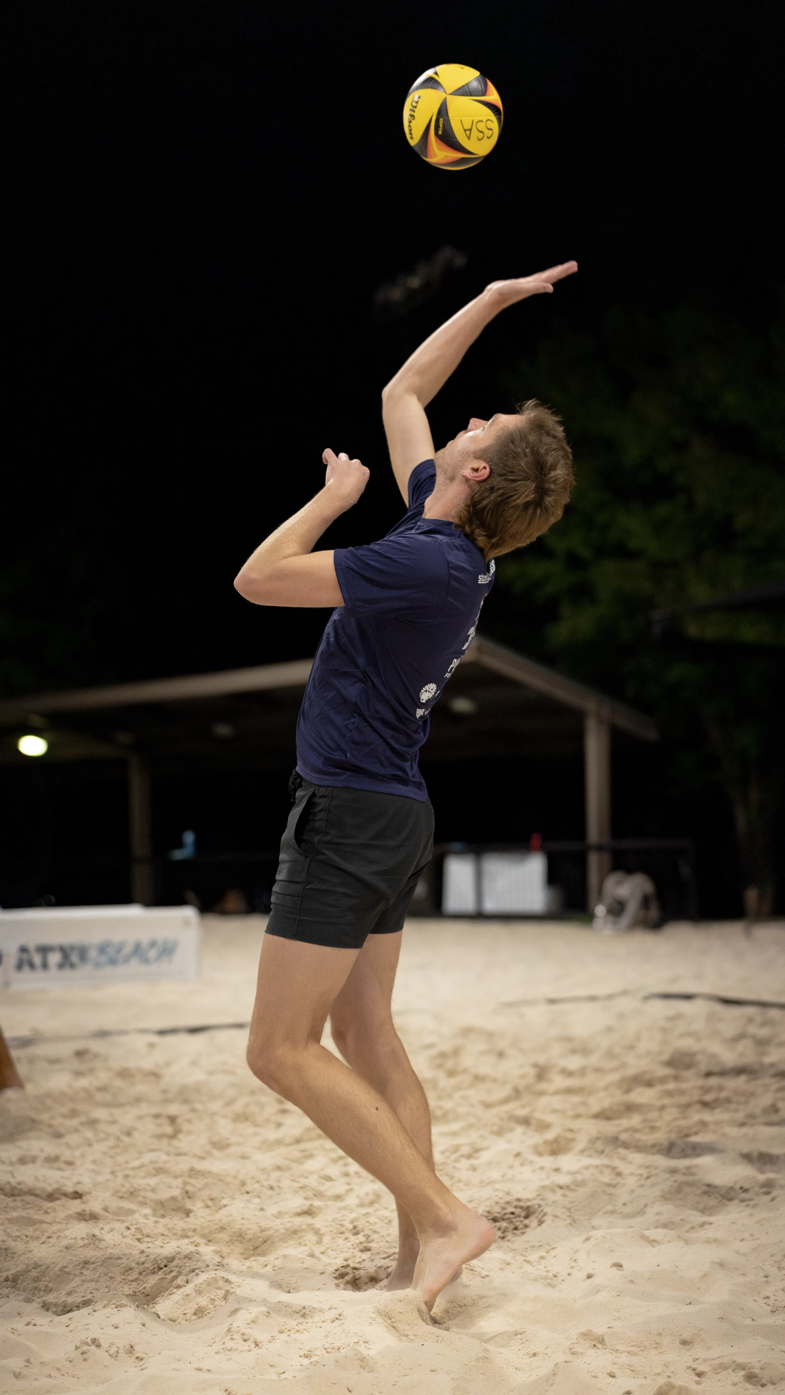 A player performs a jump serve on a lit sand volleyball court at night, tossing a yellow-and-black ball overhead and swinging through with an outstretched arm. The athlete is barefoot in the sand wearing a navy shirt and black shorts, with a pavilion and “ATX Beach” signage visible in the background.
