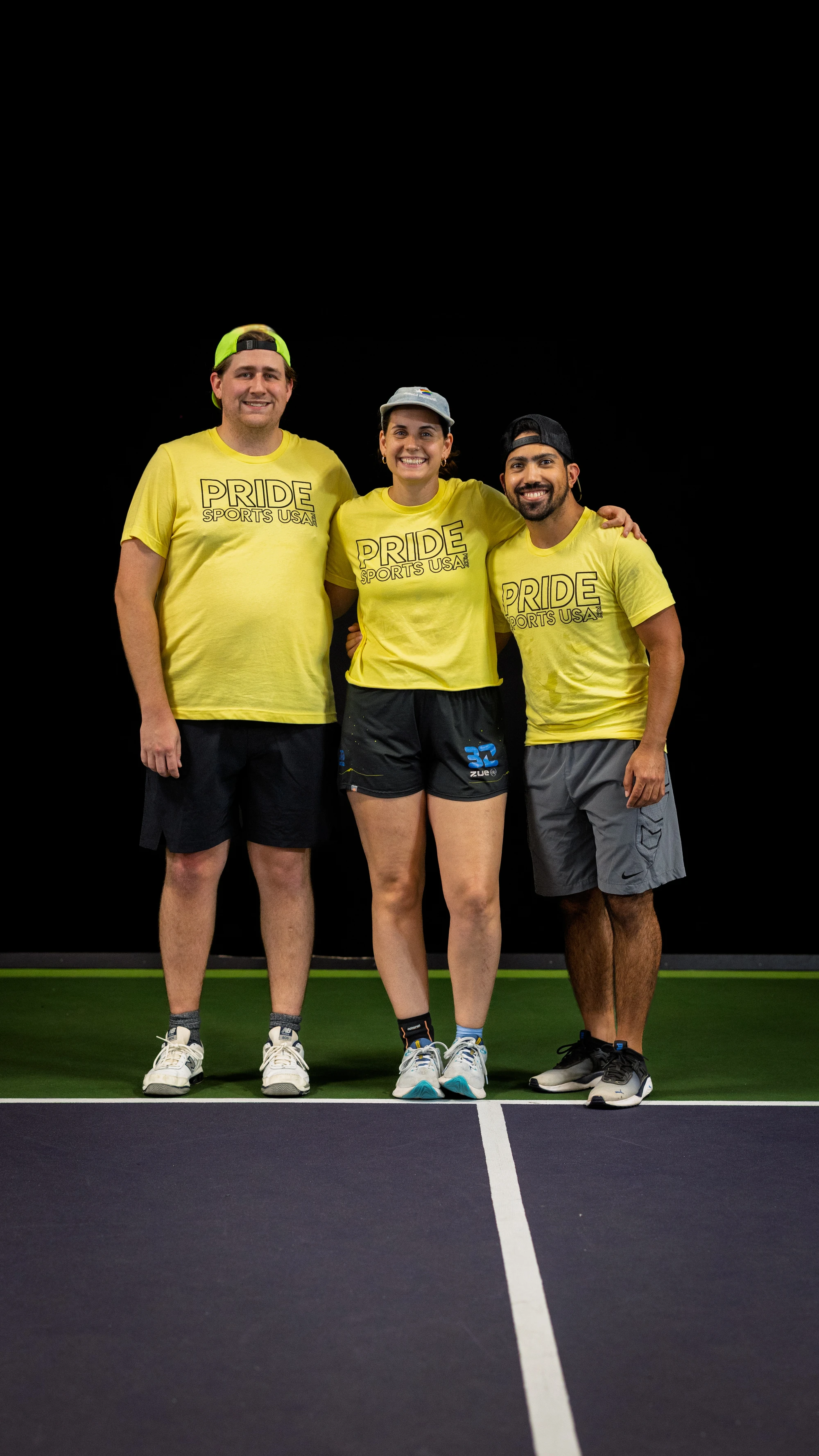 Three pickleball players stand side by side on an indoor court, each wearing matching yellow “Pride Sports USA” t-shirts, shorts, athletic shoes, and caps. The group is smiling and posing together in front of a dark background.