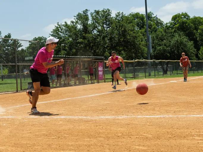A player wearing a pink shirt, black shorts, and a white cap is running toward a red kickball on a dirt field, while other players and teammates in matching pink shirts watch or move in the background near the dugout.