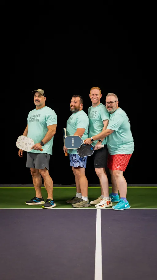 Four pickleball players stand side by side on an indoor court, each holding a paddle and wearing light blue matching "Pride Pickleball" t-shirts, athletic shorts, and sneakers. They are posing and smiling, ready for the game.