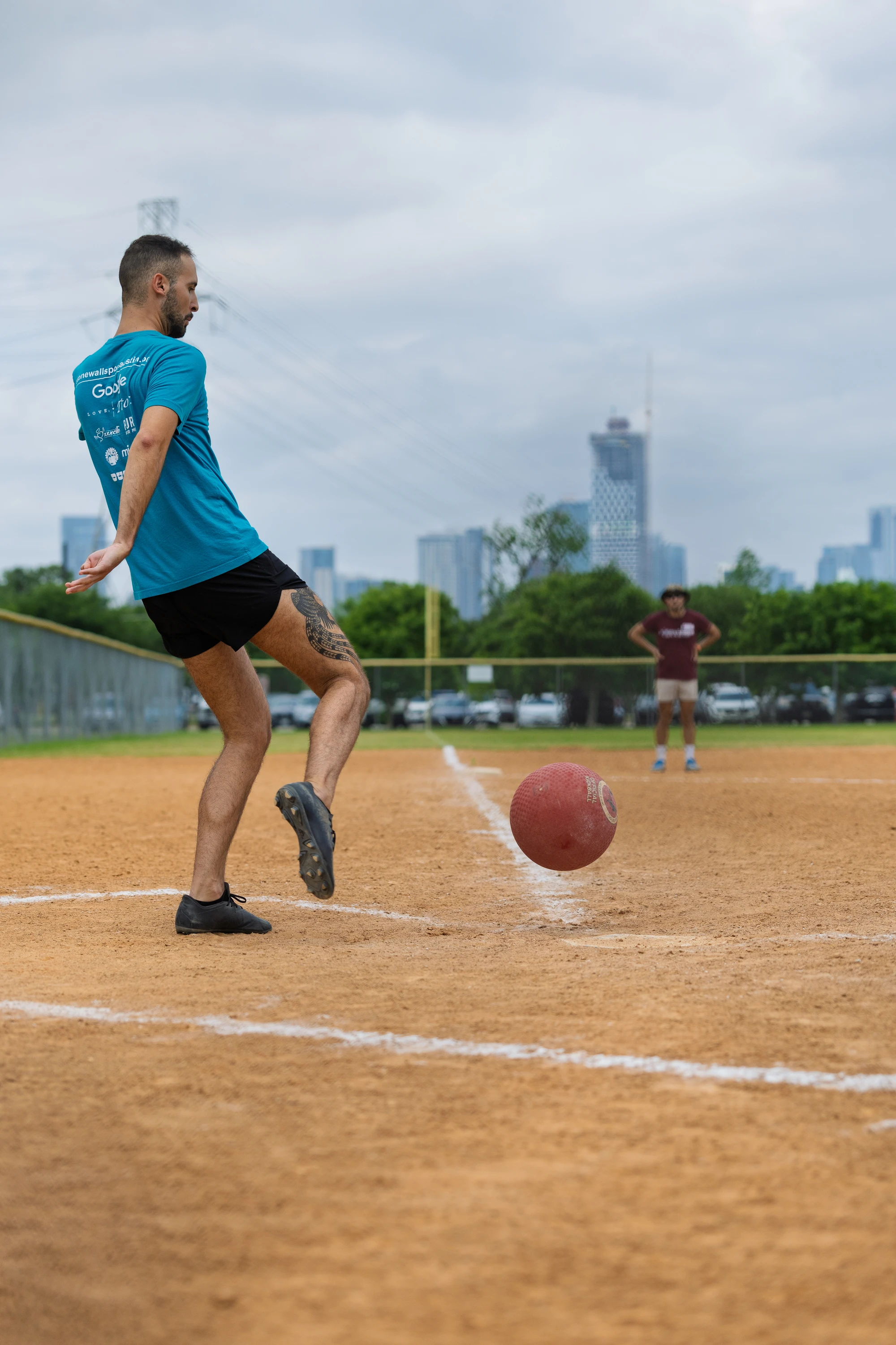 A player in a teal shirt and black shorts is kicking a red kickball on a dirt kickball field, with another player standing in the outfield and city buildings visible in the background.