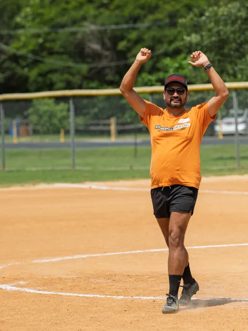 A player wearing an orange "Stonewall Kickball" shirt, black shorts, a red and black hat, and sunglasses is standing on a kickball field with both arms raised in celebration or excitement. The setting is an outdoor field with a dirt infield and green trees in the background.