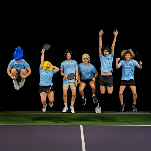 A group of pickleball players wearing colorful wigs and matching blue "Pride" shirts are on a pickleball court, energetically jumping in the air and holding their paddles.