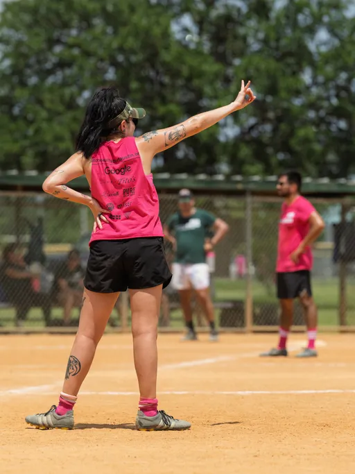 A player wearing a pink sleeveless shirt, black shorts, and pink socks stands on a kickball field, raising one arm and gesturing with two fingers while facing away from the camera; other players and a chain-link fence are visible in the background.