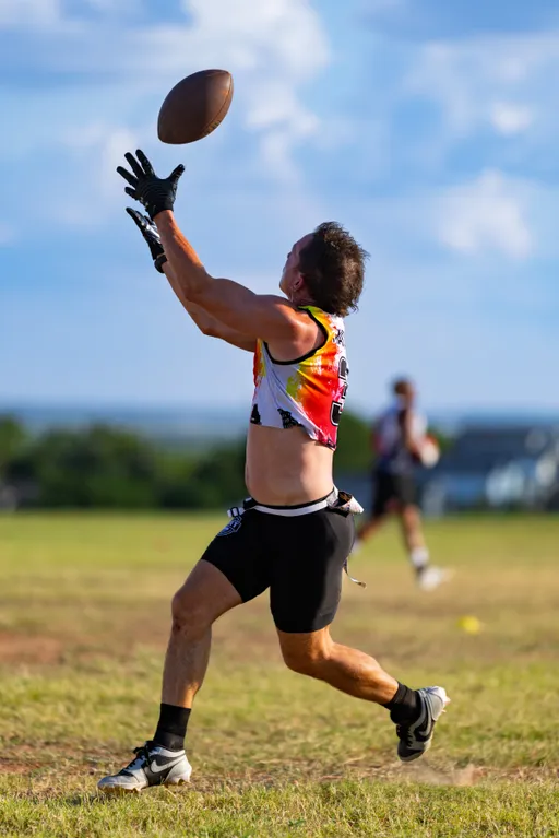 On a sunny grassy field, an ATX Heat (Black) player sprints with gloved hands raised to catch a football during flag football practice for Gay Bowl 2025. The athlete wears a multicolor sleeveless team jersey, black shorts, and cleats, with another player and cones blurred in the background.