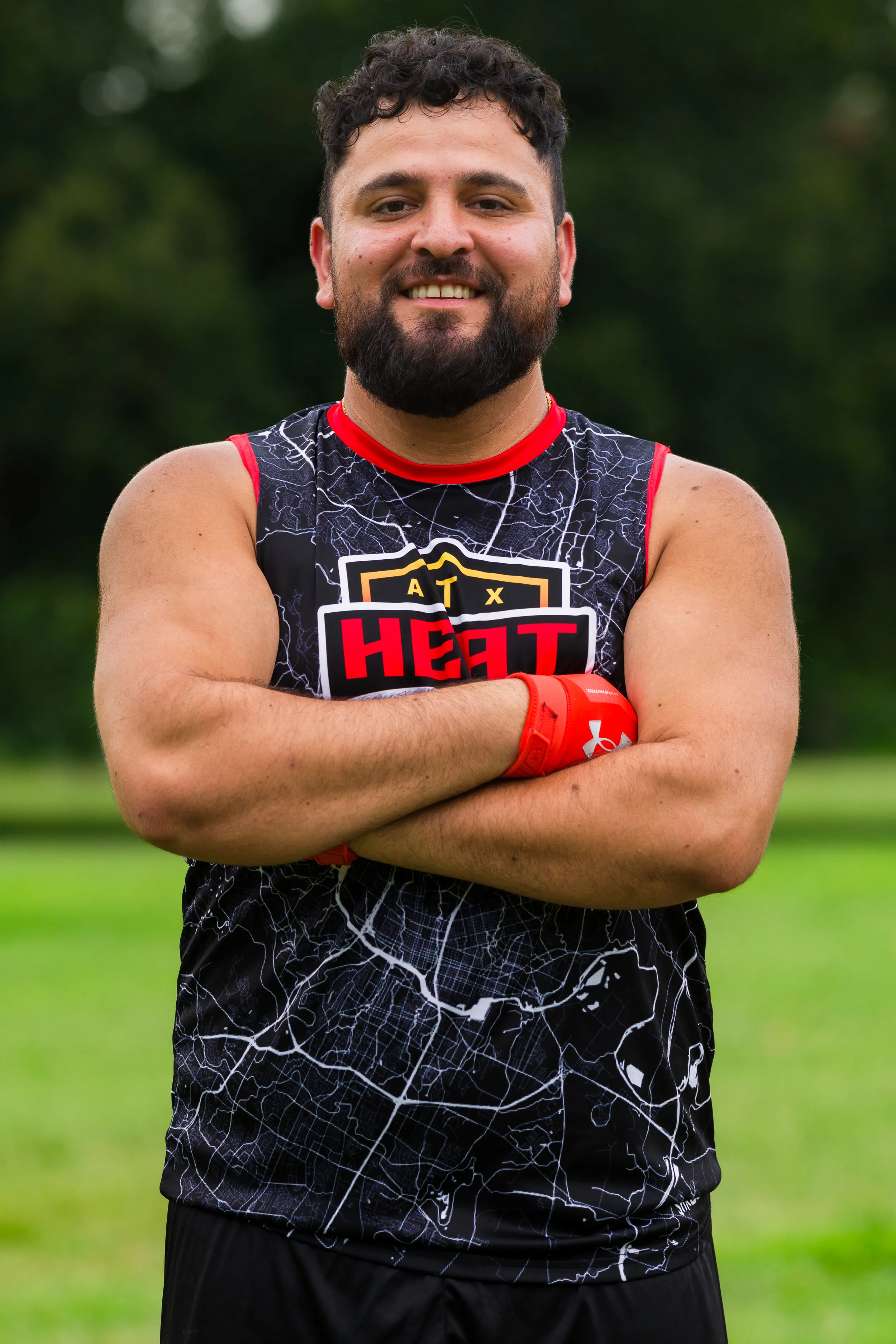 An ATX Heat (Orange) player stands on a grassy field during practice for Gay Bowl 2025, arms crossed, wearing a black map-pattern sleeveless jersey with red trim and an “ATX HEAT” logo, plus bright red receiver gloves, with trees blurred behind.