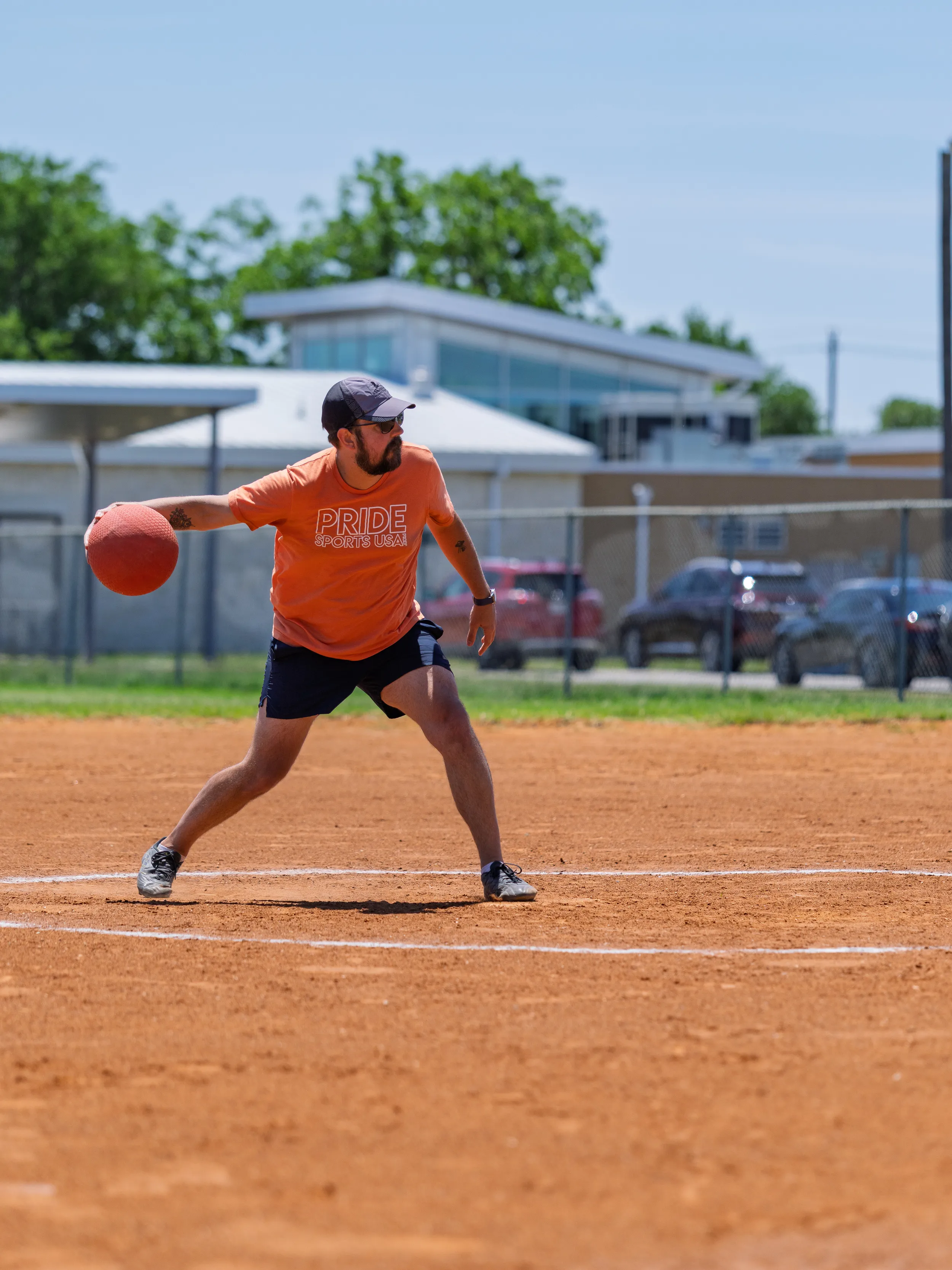 A kickball pitcher in an orange “PRIDE Sports” shirt, dark shorts, athletic shoes, and a cap winds up on the mound to roll a red kickball underhand on a sunlit dirt field, with a chain-link fence, parked cars, and low buildings in the background.