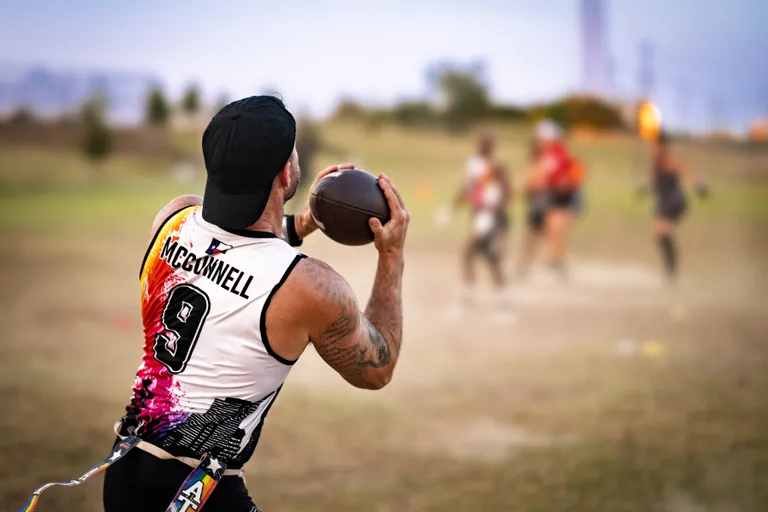 On a grassy practice field at dusk, a quarterback for ATX Heat (Black) cocks an arm to throw a football while teammates run routes downfield. The athlete wears a backward black cap, a sleeveless white jersey with a colorful graphic and the number 9, and a flag belt, with defenders and receivers blurred in the distance.