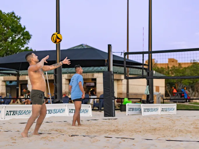 A player on a sand volleyball court is preparing to serve, with arms extended and eyes on the ball, while another player stands ready near the net; both are dressed in athletic shorts and shirts, with the court surrounded by banners and an outdoor pavilion in the background.