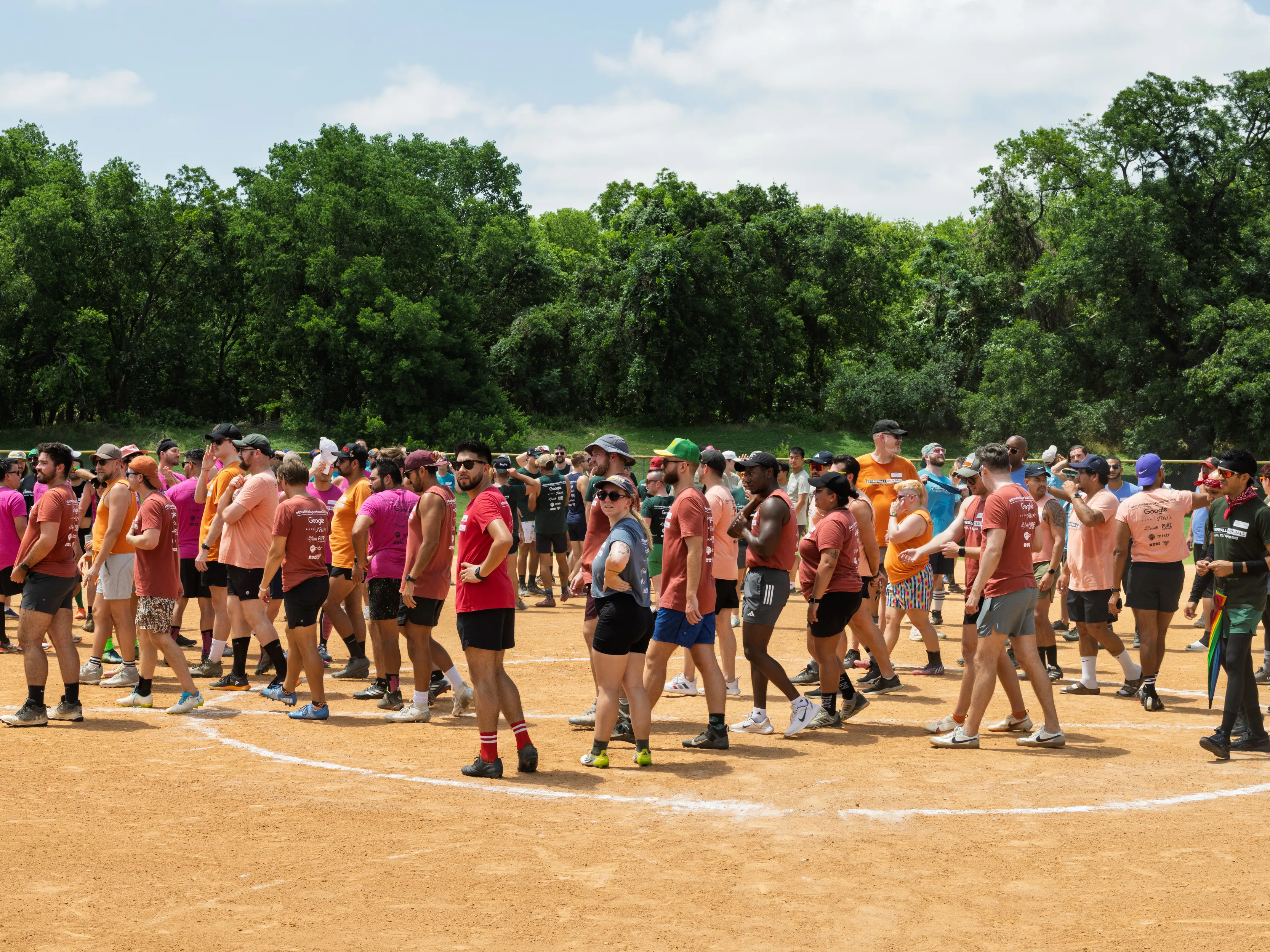 Dozens of players walk in a loose line across the infield of a kickball field, following a chalked arc, as part of a game transition or pregame activity. They wear colorful team shirts and athletic gear—caps, sunglasses, knee-high socks—while a few hold bright orange kickballs; a tree-lined park backs the sunny scene.