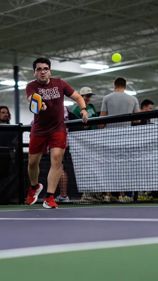 A pickleball player wearing a "Pride Sports USA" t-shirt and red shorts prepares to hit a yellow pickleball with a paddle on an indoor court, while several people watch from behind the net.
