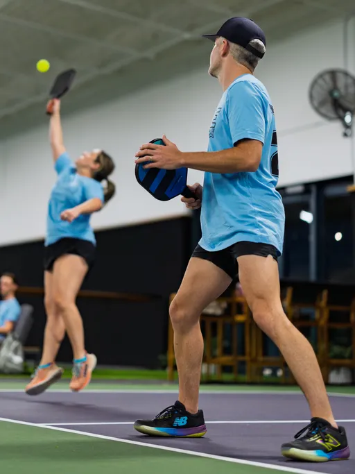 Two pickleball players in matching blue shirts and black shorts are in action on an indoor court; one player prepares to serve while the other leaps to hit the ball with a paddle.