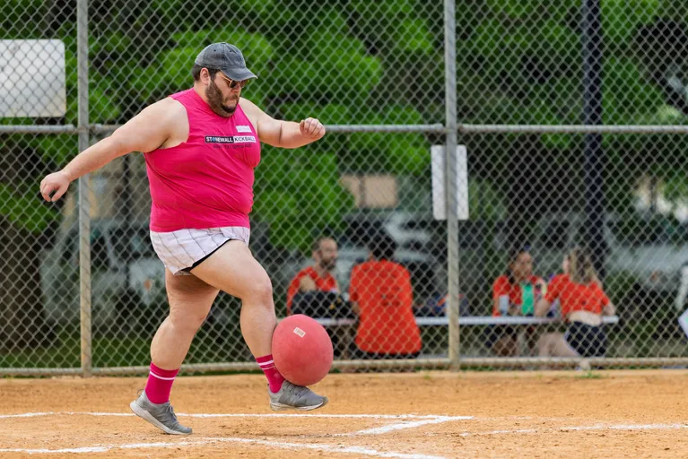 A kickball player wearing a bright pink tank top, white shorts, gray sneakers, pink socks, and a gray cap is in motion, kicking a red kickball on a dirt field while other team members watch from a bench behind the fence.