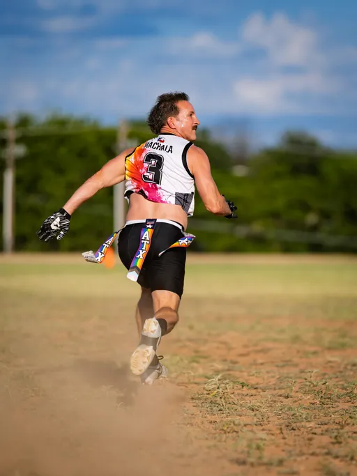 During ATX Heat (Black) practice for Gay Bowl 2025, a flag football player in a white-and-black #3 jersey, black shorts, gloves, and cleats sprints across a dusty grass field with an ATX-branded flag belt fluttering, glancing back under a bright blue sky.
