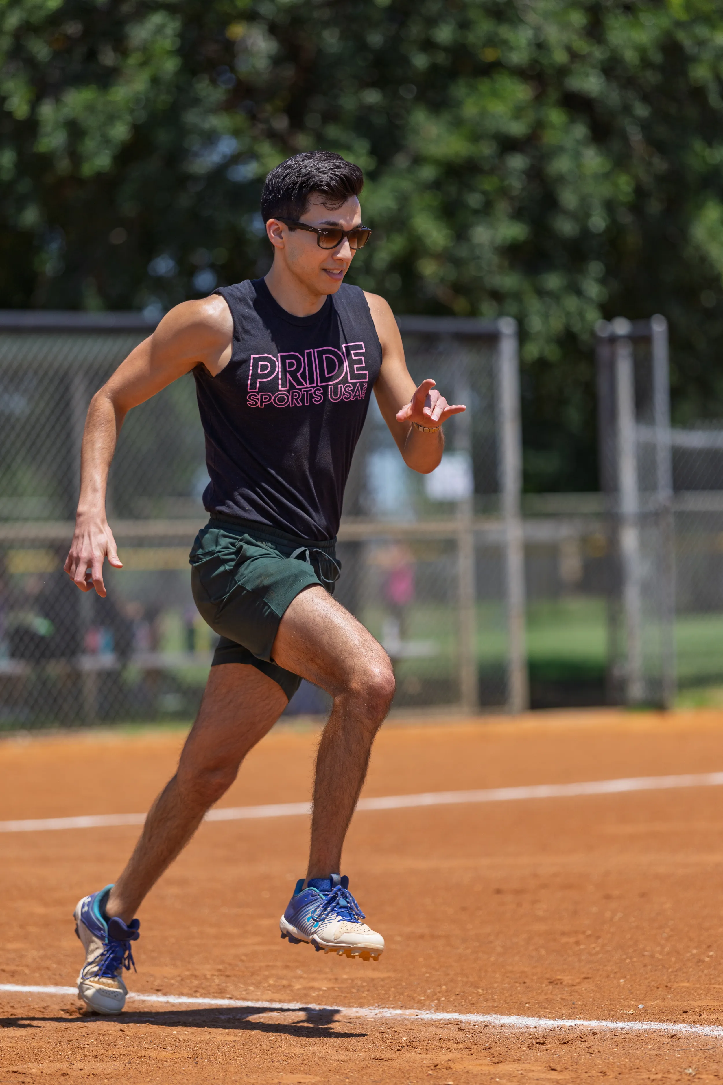 A player sprints along the chalked baseline of a sunlit kickball field, mid-stride with arms pumping, wearing a black tank top reading “PRIDE SPORTS USA,” dark green shorts, blue-and-white running shoes, and sunglasses; chain-link fences and trees line the background.