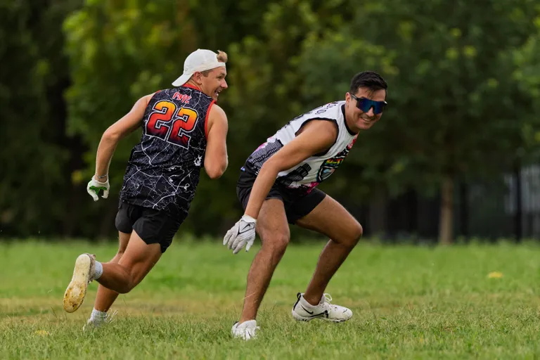 Two ATX Heat (Orange) athletes run a flag football drill on a grassy field, with one in a black jersey showing number 22 and a backward cap sprinting after a teammate in a white jersey and mirrored sport sunglasses who cuts to change direction; both wear cleats and gloves, with trees soft in the background.