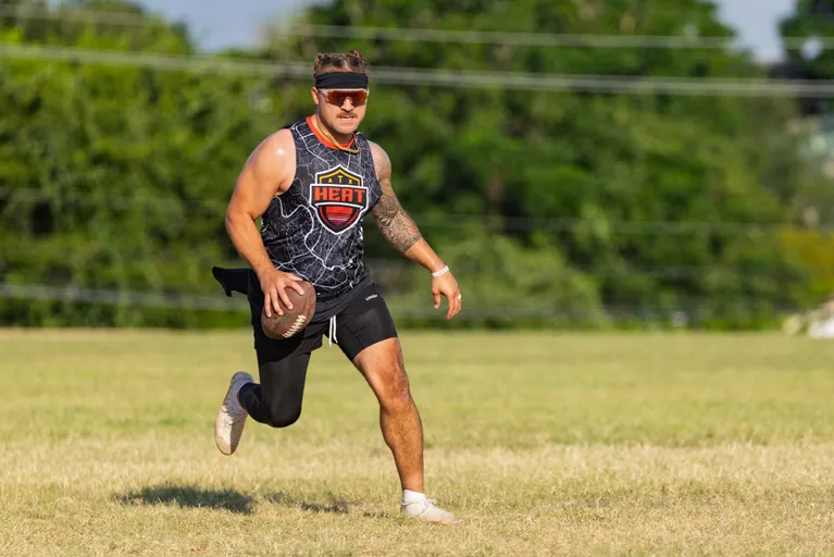 An ATX Heat (Black) player runs across a grassy field during Gay Bowl 2025 practice, cradling a football. The athlete wears a black patterned sleeveless jersey with the ATX Heat crest, black shorts, white shoes, a headband, sport sunglasses, and a flag belt, with trees in the background under bright sun.