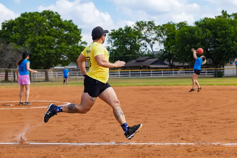 On a sunny kickball field, a player in a yellow jersey, black shorts, patterned socks, and a black cap sprints up the baseline as chalk dust trails behind. A blue-shirt fielder at first base reaches to catch the red kickball while other players in blue wait in the infield, with trees and a fence in the background.