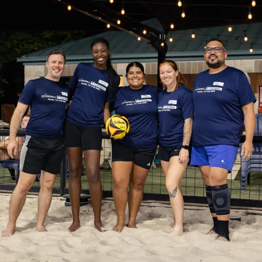 Five players pose together on a sand volleyball court under string lights, wearing matching navy Stonewall Sand Volleyball shirts; the center player holds a yellow Wilson volleyball. A fenced court with a gazebo and benches is in the background, and one player wears a knee brace with everyone in athletic shorts.