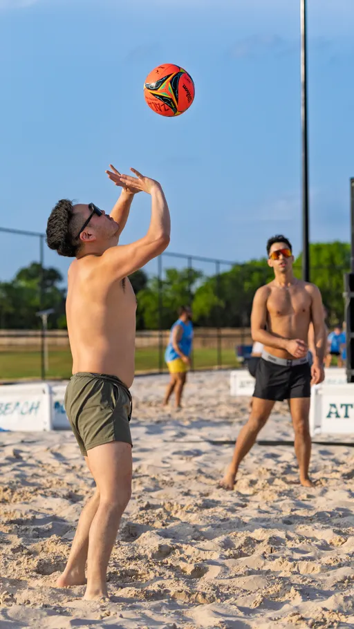 A player wearing shorts and sunglasses prepares to set a volleyball underhand as another competitor, also in shorts and sunglasses, stands ready nearby on a sandy outdoor volleyball court.