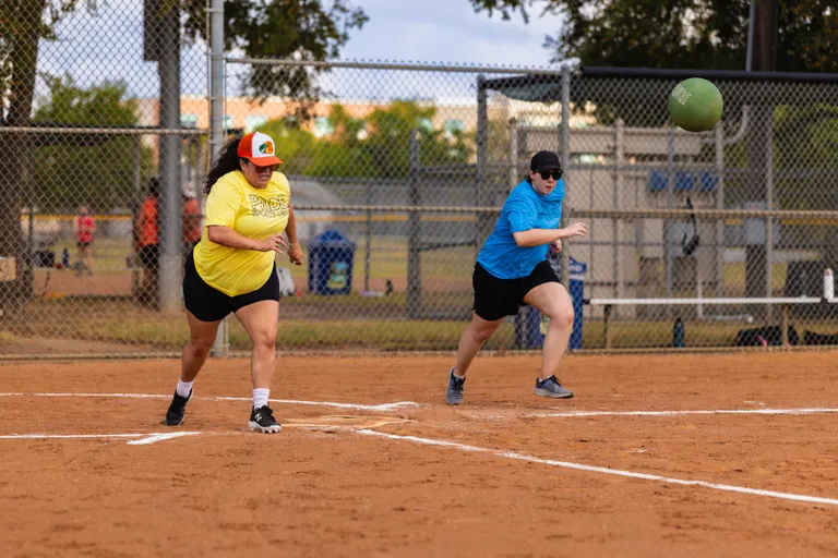 On a dirt kickball field, a player in a yellow Pride Kickball shirt and multicolor cap sprints from home plate after a kick while a defender in a blue team shirt, black cap, and sunglasses charges toward a green kickball; white baselines, chain-link fencing, and trees frame the park setting.