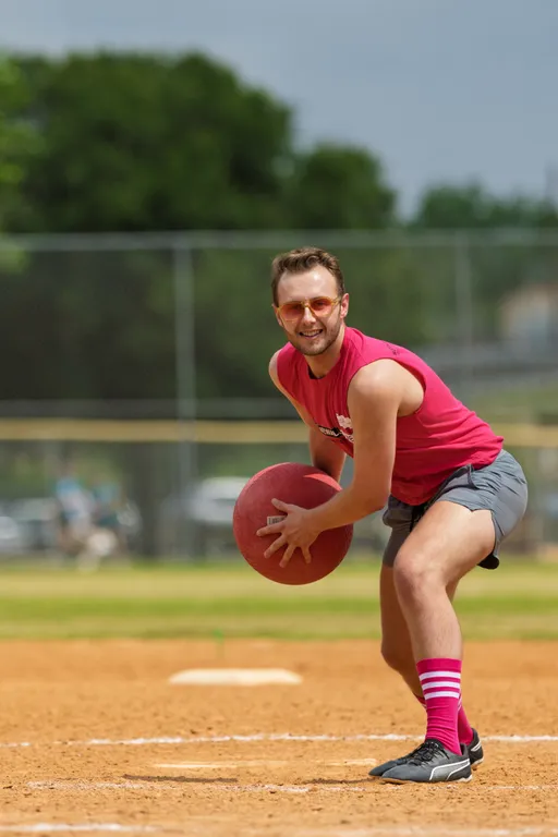 A player wearing a sleeveless red shirt, gray shorts, pink-striped knee socks, and athletic shoes is preparing to pitch a red kickball on a dirt kickball field, with green trees and a chain-link fence in the background.