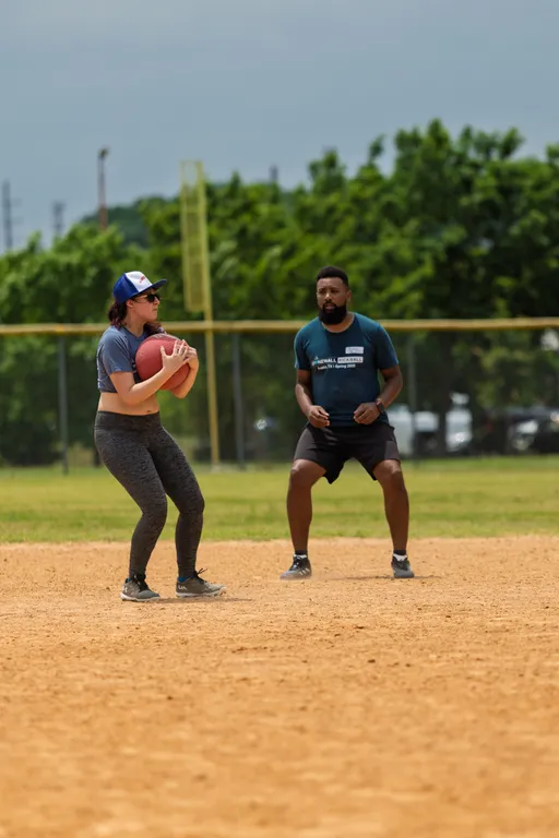 Two kickball players are on a dirt field; one player in a blue cap and athletic clothing is holding a red kickball, while another player in a dark shirt and shorts appears ready to move or defend, with a grassy outfield and trees in the background.