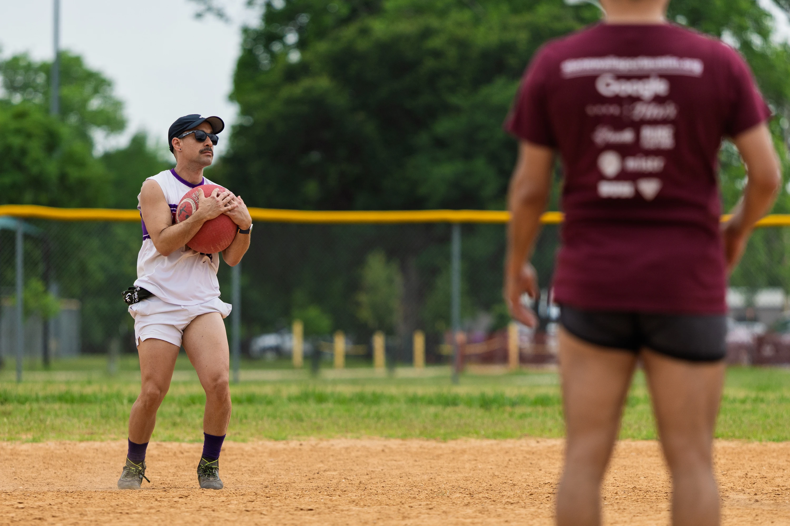 A kickball player in a white uniform, hat, and sunglasses holds a red kickball while preparing to throw, as another player in a maroon shirt and black shorts stands in the foreground on a dirt field with a green outfield and trees in the background.