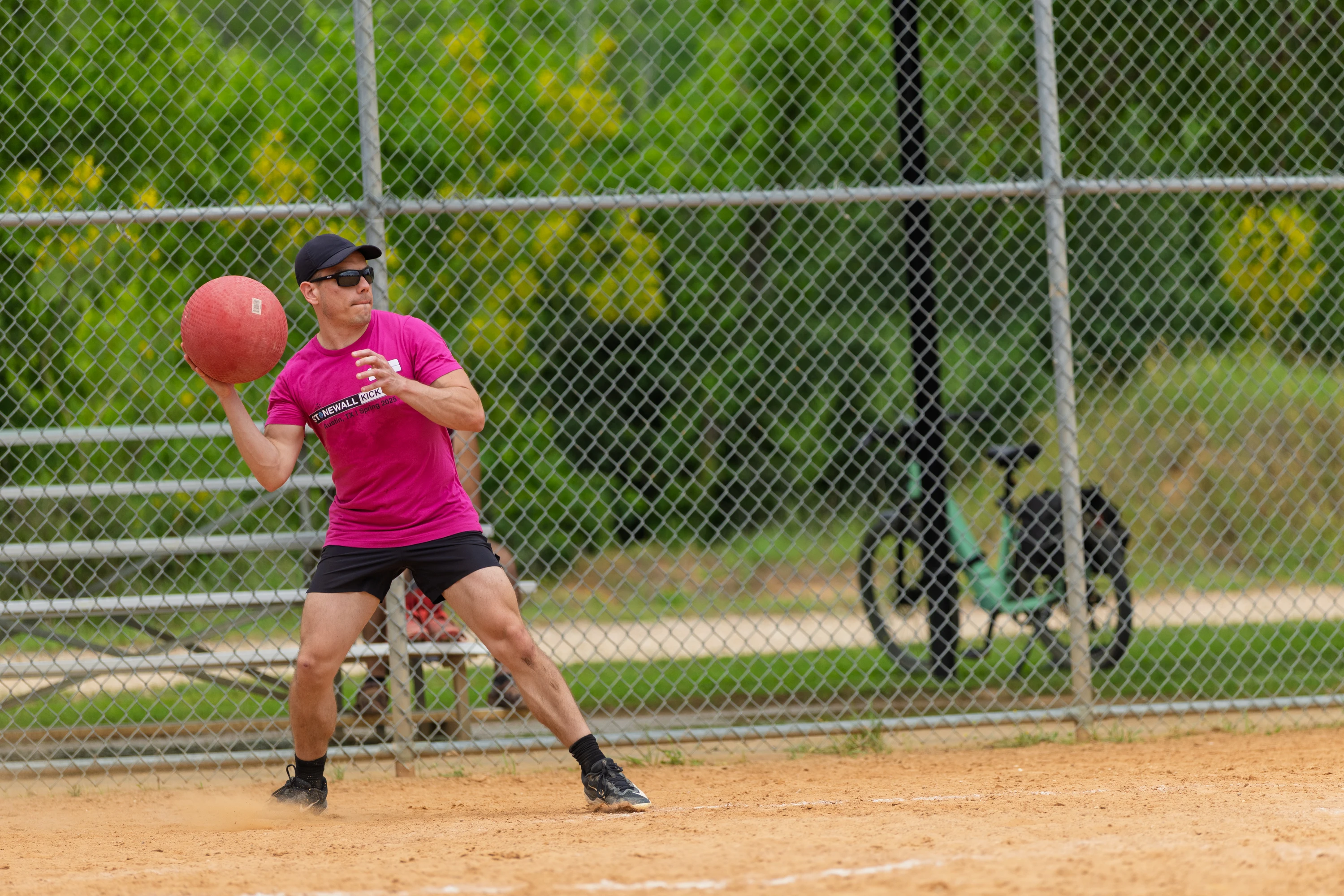 A kickball player wearing a bright pink shirt, black shorts, a black cap, and sunglasses is preparing to throw a red kickball on a sandy field, with a chain-link fence, green trees, and bicycles visible in the background.