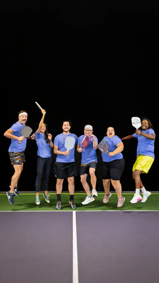 Six pickleball players wearing matching blue shirts pose in mid-jump on an indoor pickleball court, each holding a paddle and appearing enthusiastic and celebratory.