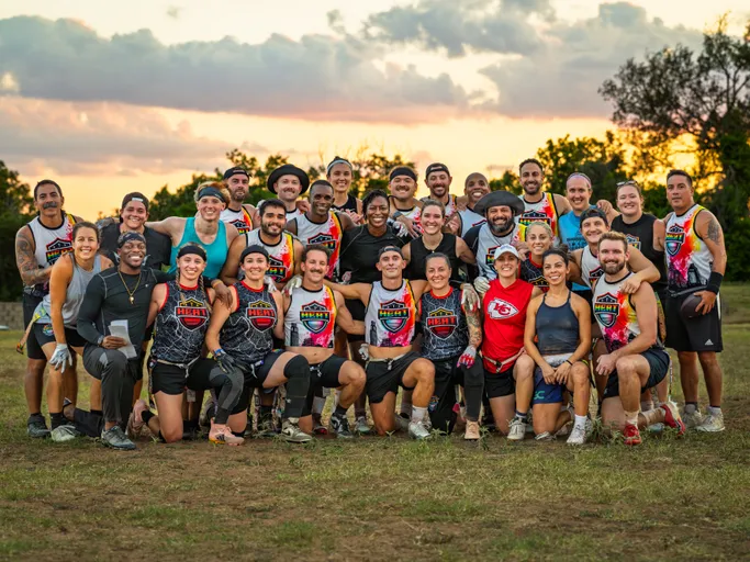 A large group of ATX Heat (Black) flag football players pose together on a grassy field at sunset during Gay Bowl 2025 prep. Athletes wear ATX Heat jerseys—black, white, and rainbow-accented—plus shorts and cleats; some sport gloves, visors, headbands, and knee sleeves.