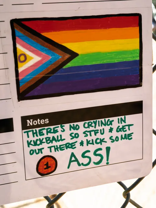 A sign attached to a chain link fence displays a colorful Progress Pride flag and a handwritten motivational message encouraging players to be tough and energetic during the kickball game.