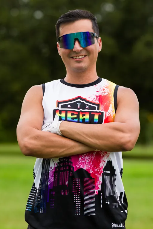 An ATX Heat player stands on a grassy practice field with arms crossed, wearing a sleeveless ATX Heat jersey with a colorful skyline and orange accent, reflective wraparound visor sunglasses, and a white football glove. Trees blur in the background, suggesting a relaxed portrait during Gay Bowl practice.